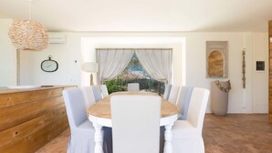 Un comedor con una mesa redonda de madera, sillas blancas tapizadas, una lámpara colgante de fibra natural, un reloj en la pared y vistas al verdor a través de una ventana con cortinas translúcidas.