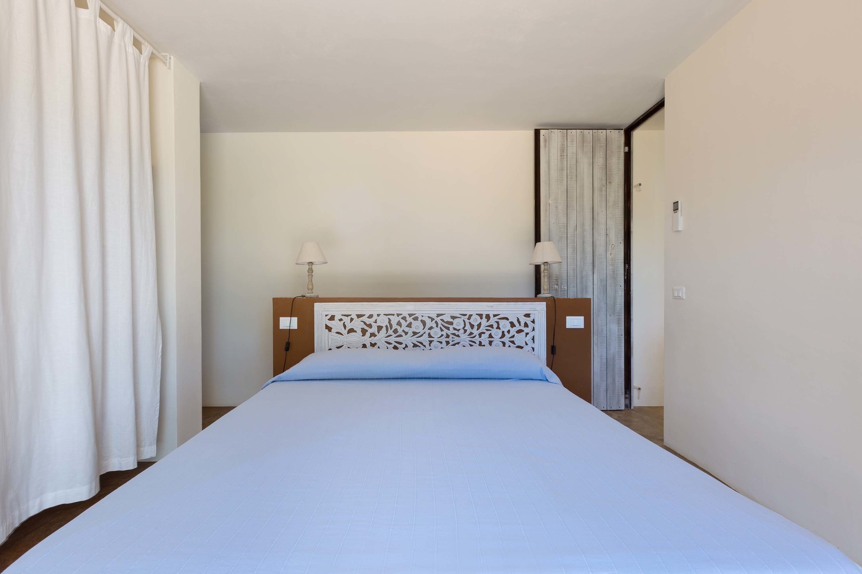 A bedroom with a bed, curtains, a nightstand with a lamp, and a door.
