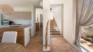 Una cocina moderna con armarios de madera, una zona de comedor con una mesa redonda, una lámpara de pie alta y una escalera que sube a otro nivel.