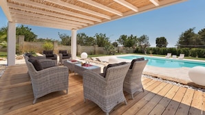 Un espacio exterior techado con mobiliario de mimbre, una piscina y vistas al paisaje circundante.