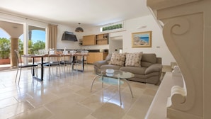 Amplia sala de estar con una mesa de centro de vidrio, un sofá y una mesa de comedor con sillas. Se observa una zona de cocina con armarios y una ventana.