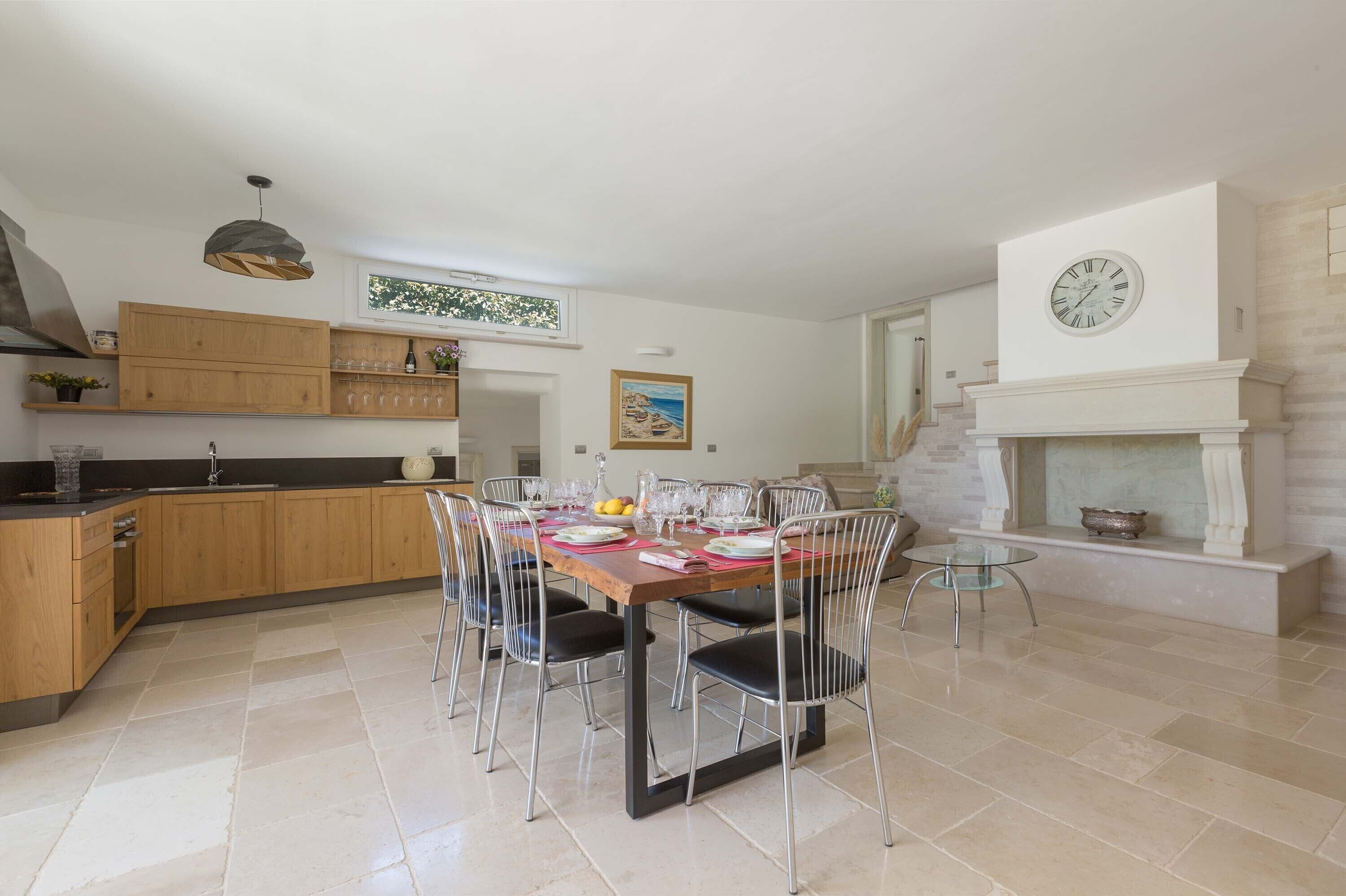 A modern kitchen with wooden cabinets, a dining table set for six, and a fireplace with a clock above it.