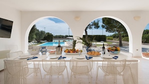 A dining area with a view of a pool, white furniture, and a table set with plates, cutlery, and a fruit bowl. - Villa Maria Clara (Gagliano del Capo)