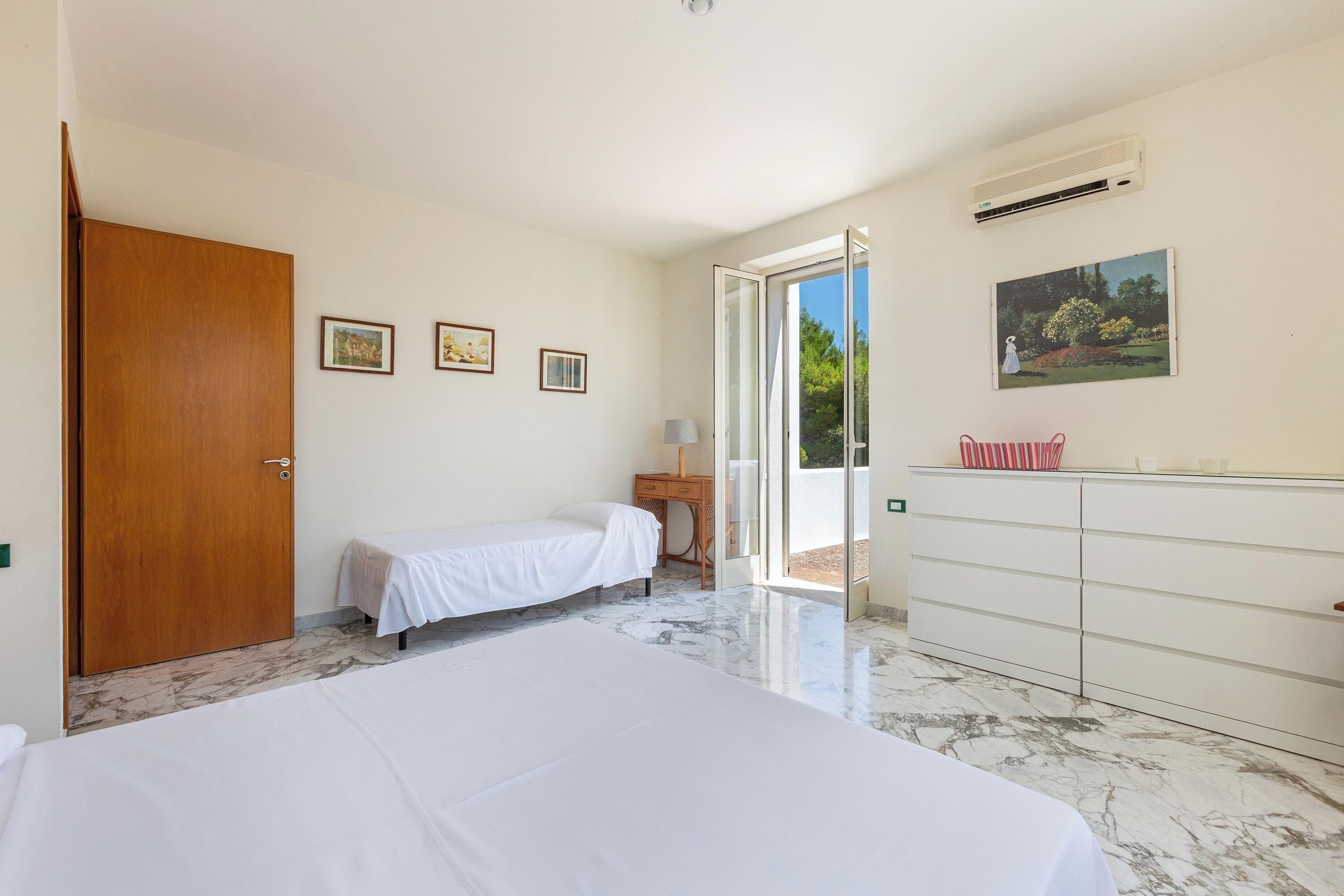 A bedroom with two beds, a wooden door, a dresser, a small table with a lamp, and a picture on the wall.