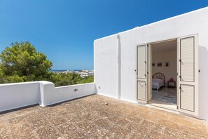 A balcony with a stone floor, a white wall, and a bedroom with a bed and a nightstand.