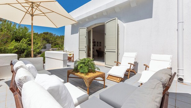 Un espace extérieur ensoleillé, aménagé avec des chaises blanches dotées de coussins, une table en osier agrémentée d’une plante et des parasols de couleur beige.
