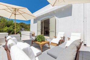 A sunlit outdoor seating area with white cushioned chairs, a wicker table with a plant, and beige umbrellas. - Villa Merienne (Castrignano del Capo)