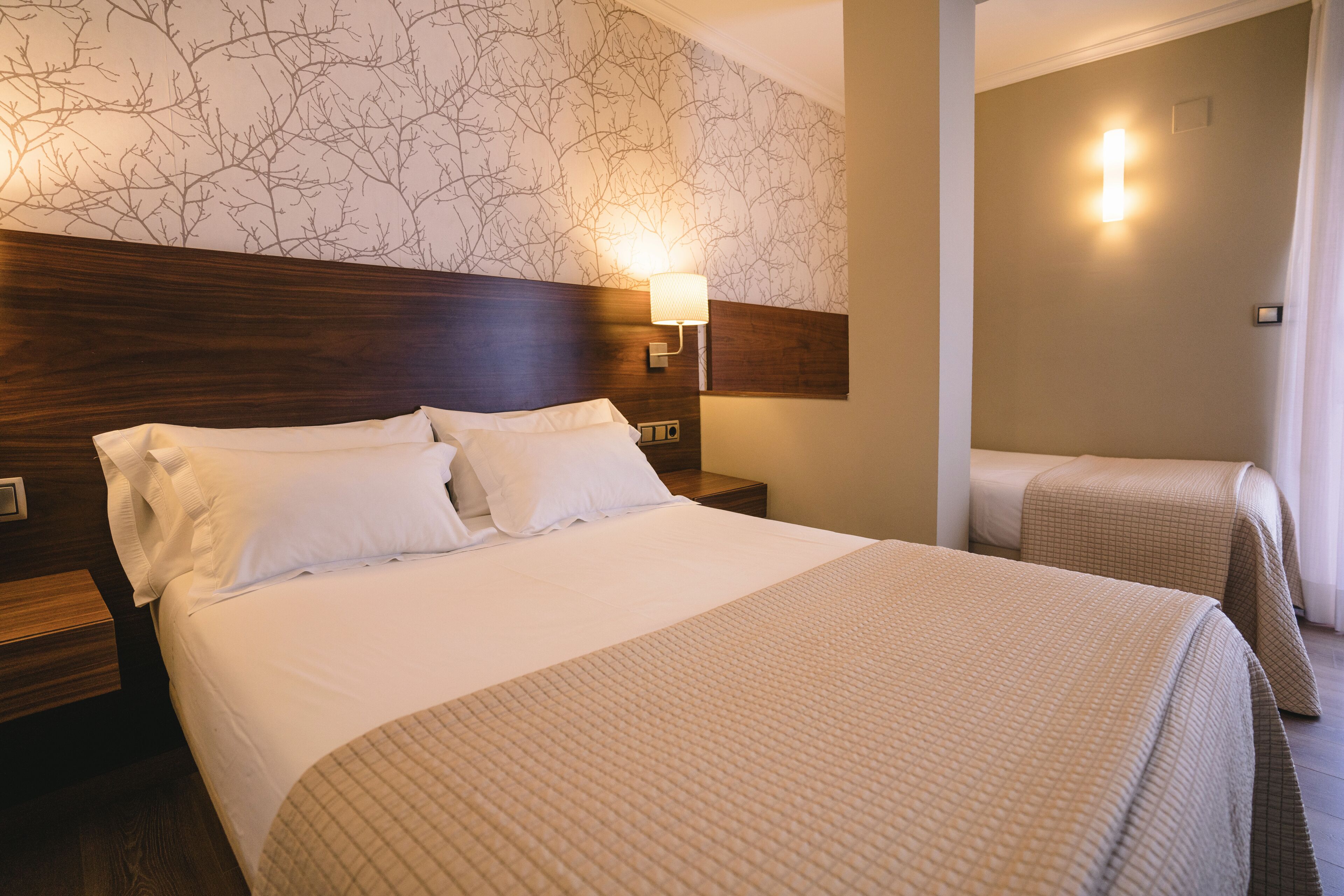 Quarto de hotel com uma cama grande, uma cama menor e uma mesa de cabeceira com abajur.