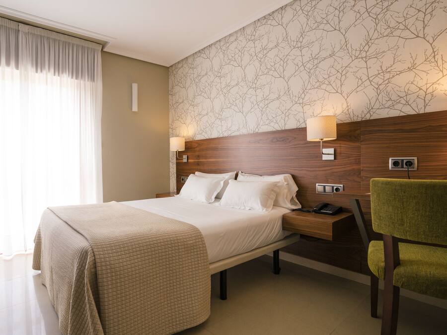 A hotel room with a large bed, bedside tables, a chair, and a patterned wallpaper.