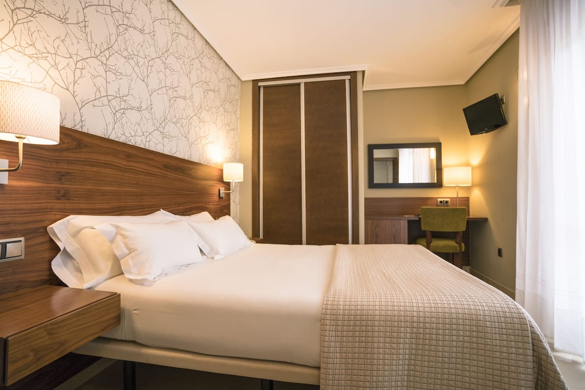 Habitación de hotel con cama, mesita de noche, lámpara, espejo y televisor montado en la pared.