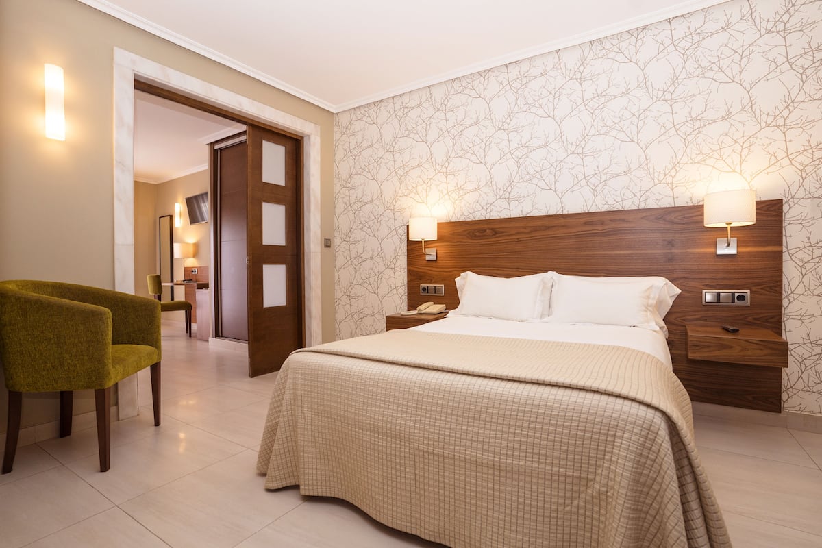 Una habitación de hotel con una cama grande, dos lámparas de noche, un cabecero de madera, un sillón verde y una puerta que da a un baño.