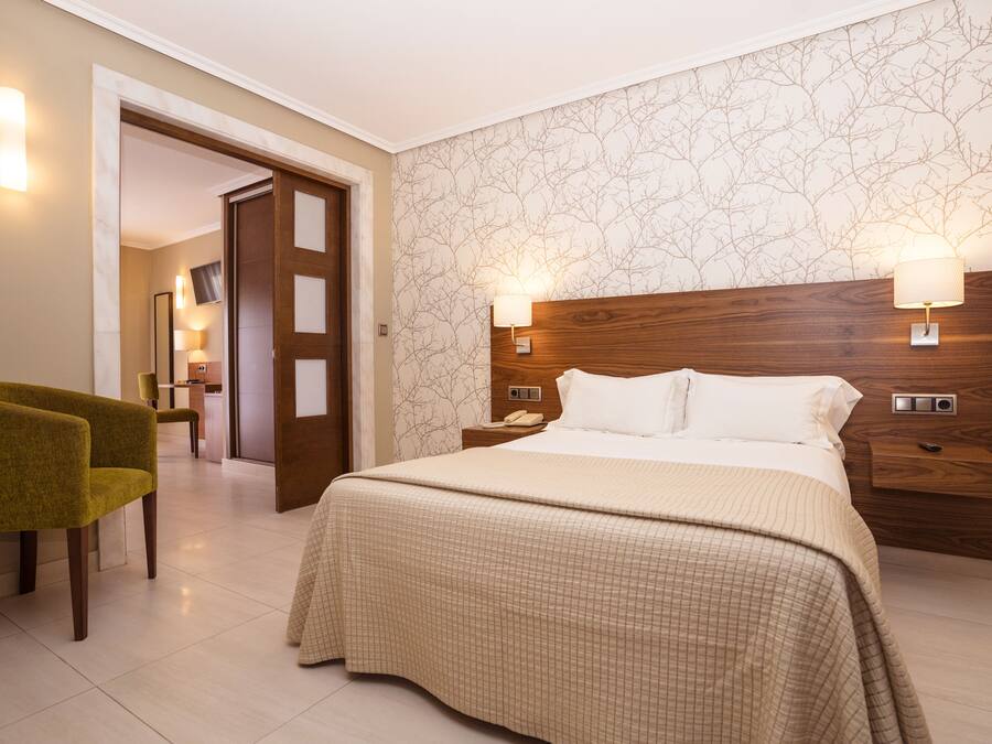 A hotel room with a large bed, two bedside lamps, a wooden headboard, a green armchair, and a door leading to a bathroom.