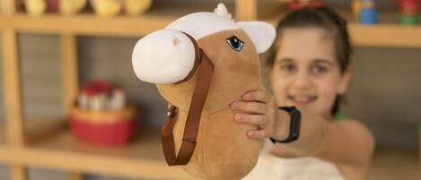 A child holding a horse head toy in front of a shelf with various toys and a 'WELCOME' sign.