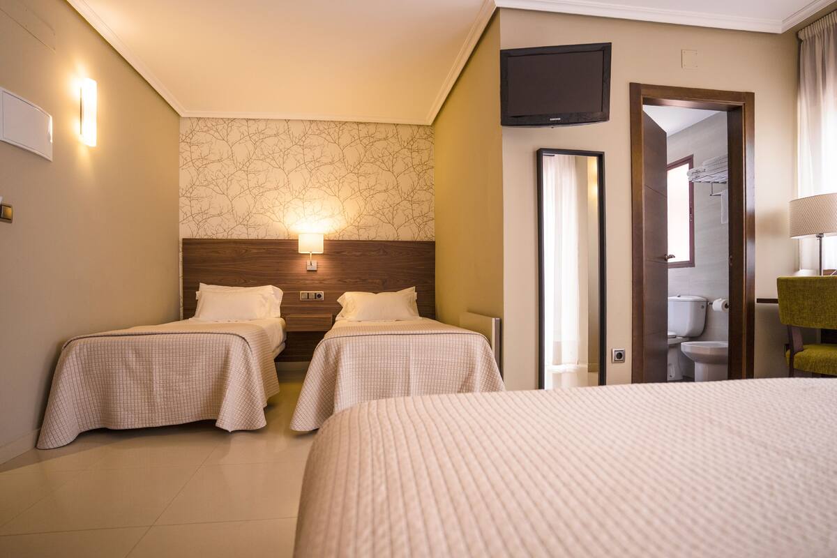 Habitación de hotel con dos camas individuales, una cama grande, cabecero de madera, televisor montado en la pared y un baño visible a través de una puerta abierta.