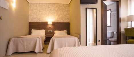 A hotel room with two single beds, a large bed, a wooden headboard, a wall-mounted TV, and a bathroom visible through an open door.