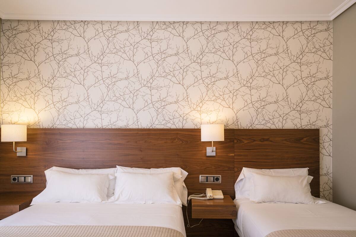 Habitación de hotel con dos camas, cabeceros de madera, una pared con diseño de ramas de árbol y lámparas de noche.