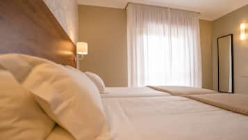 Ein Hotelzimmer mit zwei Betten, einem Holzkopfende, einem Nachttisch mit Lampe, einem großen Fenster mit leichtem Vorhang und einem Spiegel an der Wand.