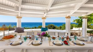 Mesa lista para comer con vista al mar, techo de madera blanca y zona de estar al aire libre.