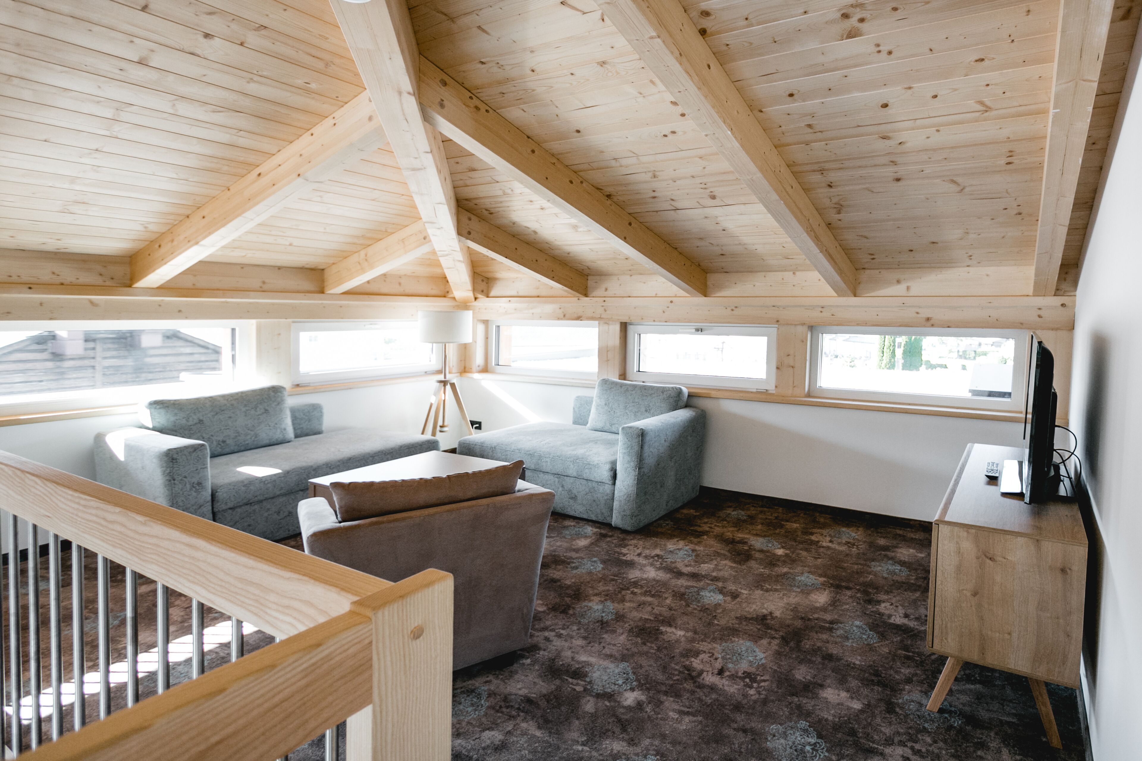 Une pièce spacieuse avec un plafond haut, des poutres apparentes en bois, un canapé gris, une petite table et une lampe.