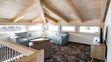 Une pièce spacieuse avec un plafond haut, des poutres apparentes en bois, un canapé gris, une petite table et une lampe.