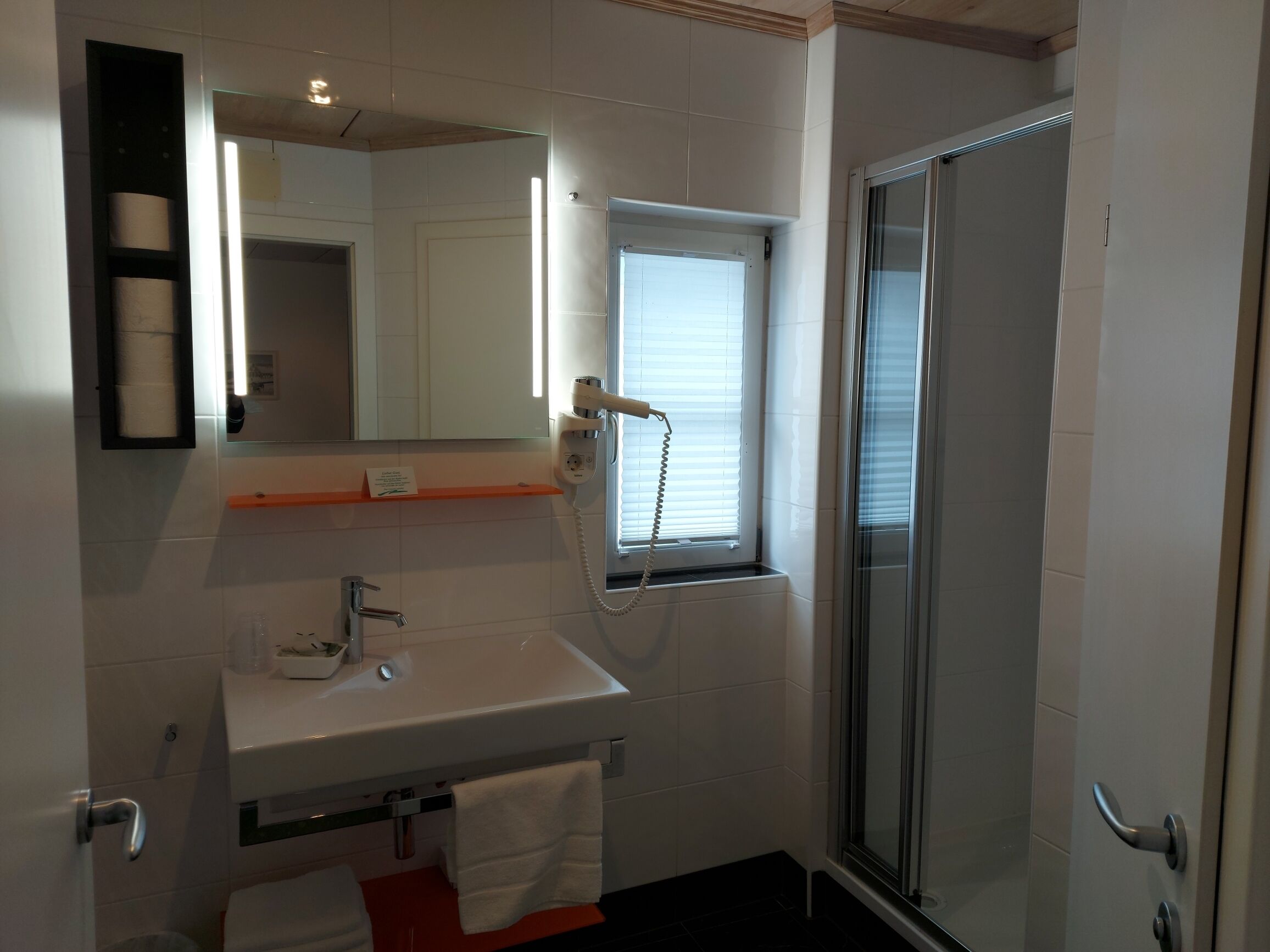 Un baño con un lavamanos blanco, un espejo, un perchero para toallas, una ducha con puerta de cristal y una ventana con persianas.
