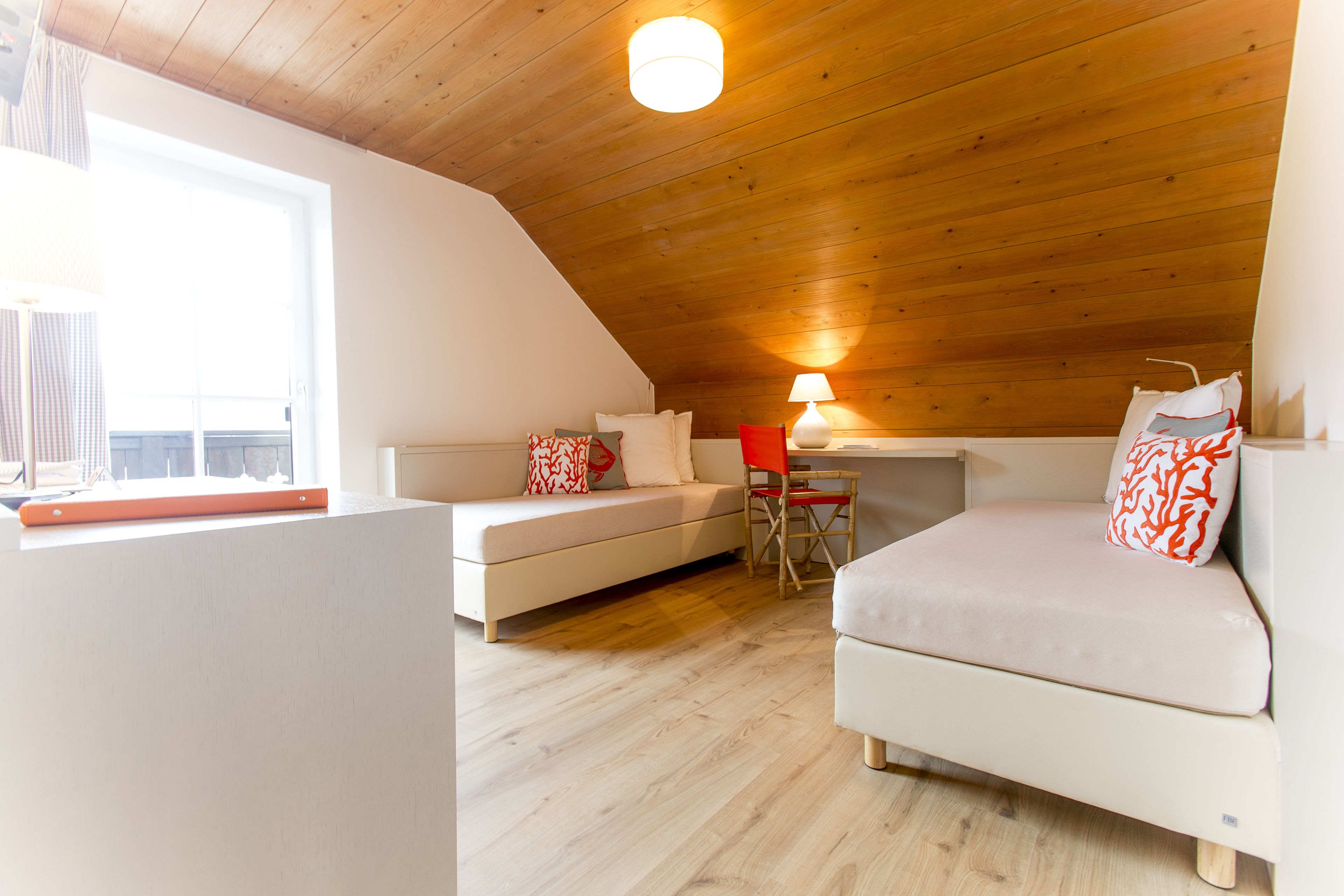 A cozy loft bedroom with a sloped ceiling, a white sofa, a red chair, and a desk with a lamp.
