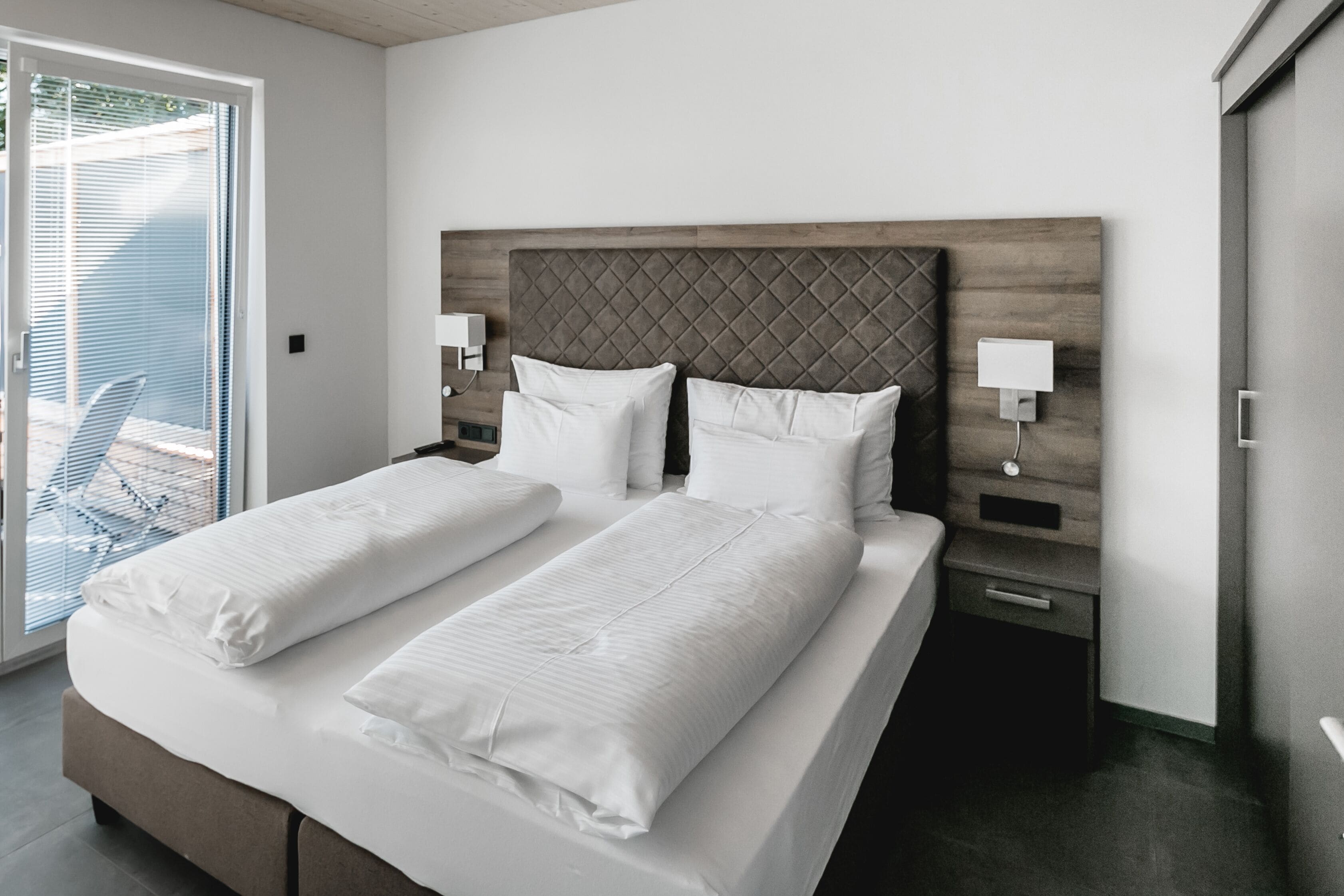 A double bed with white linens, a wooden headboard, bedside tables with lamps, and a sliding glass door to a balcony.
