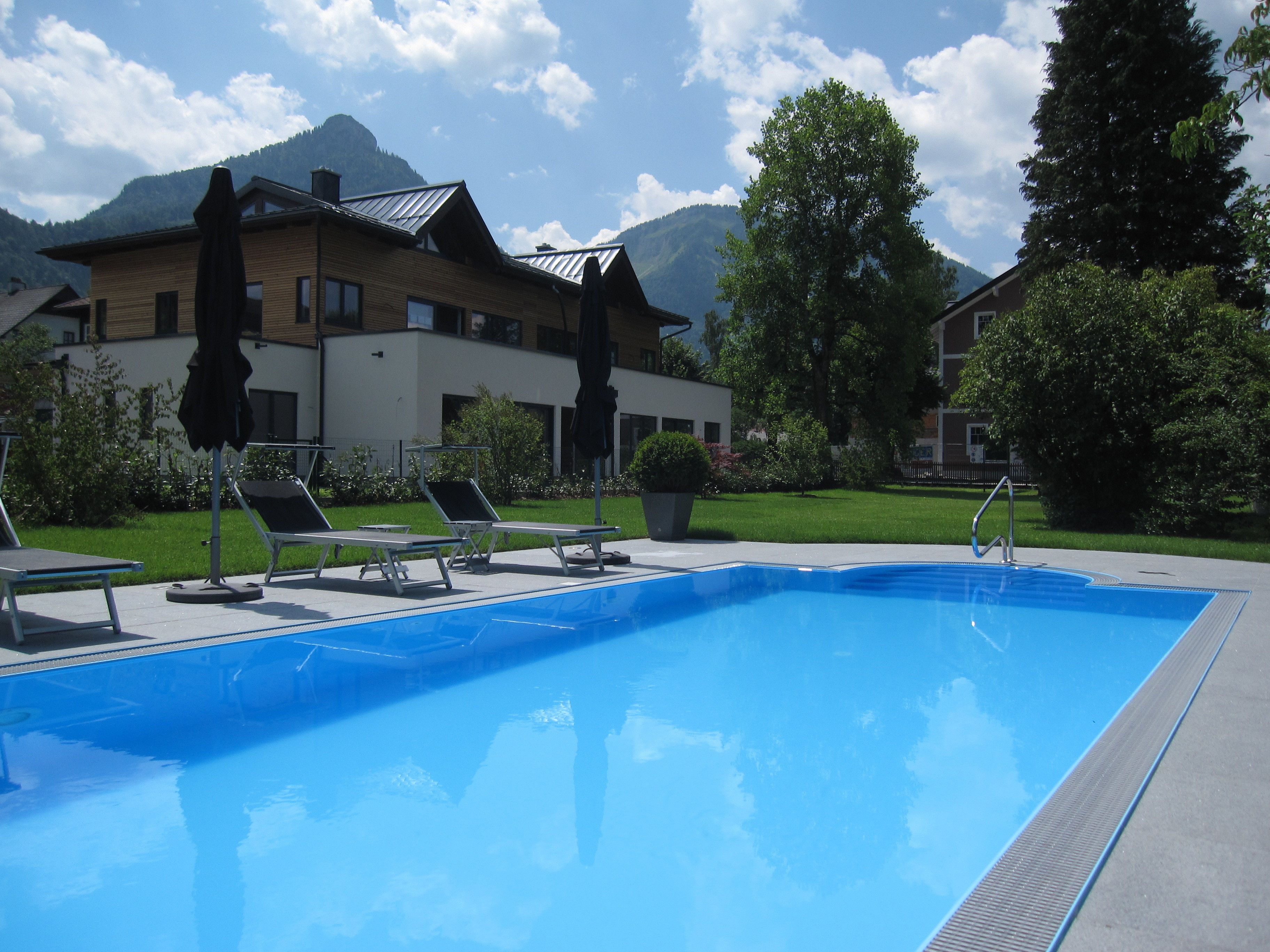A swimming pool with lounge chairs and umbrellas, surrounded by greenery and buildings with a mountainous backdrop.