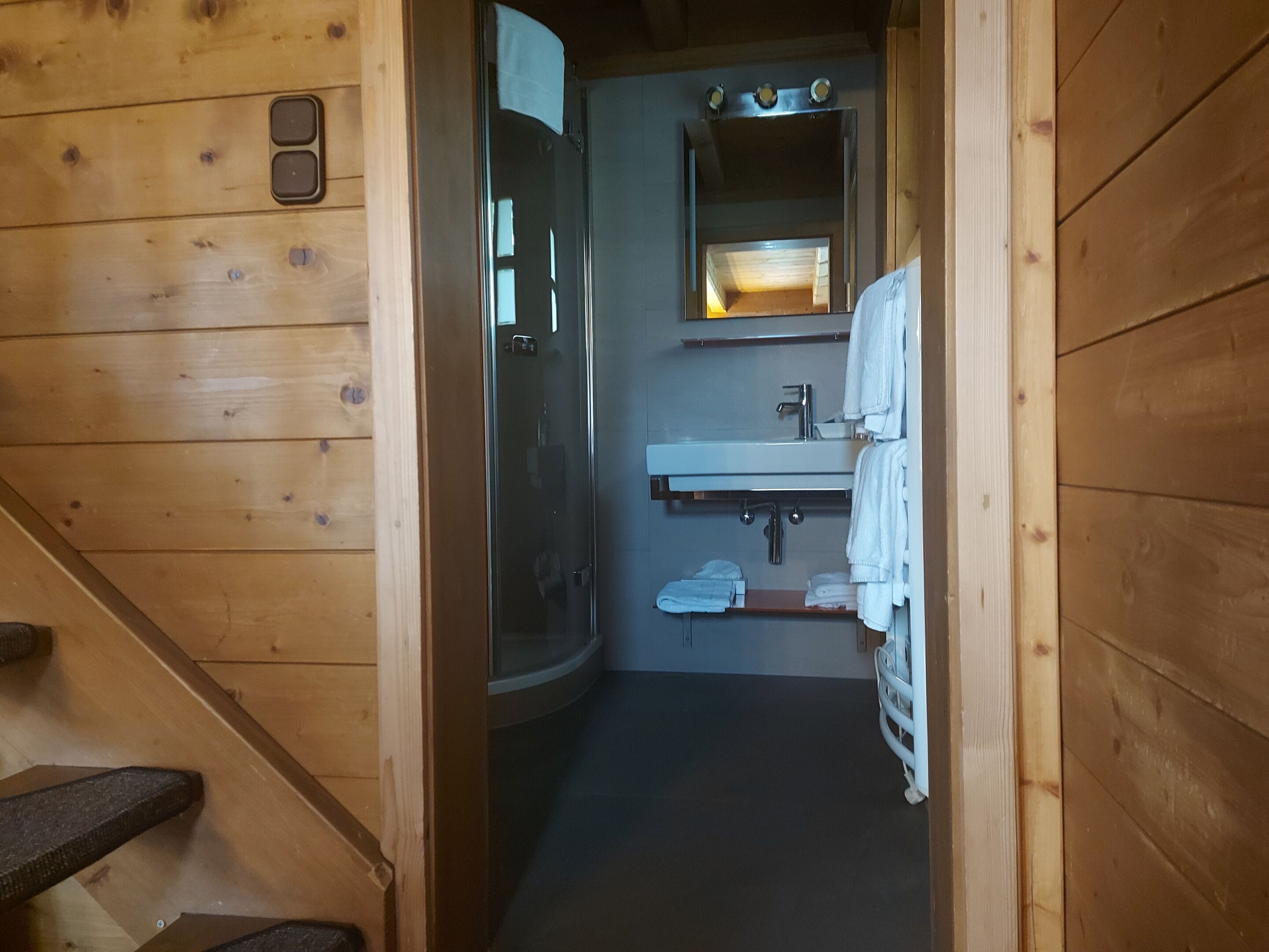 Baño de cabaña de madera con ducha de vidrio, lavamanos con espejo y toallero.