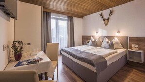 A hotel room with a bed, bedside tables, a desk with a chair, a potted plant, and a window with curtains. - Panorama (Finkenberg)