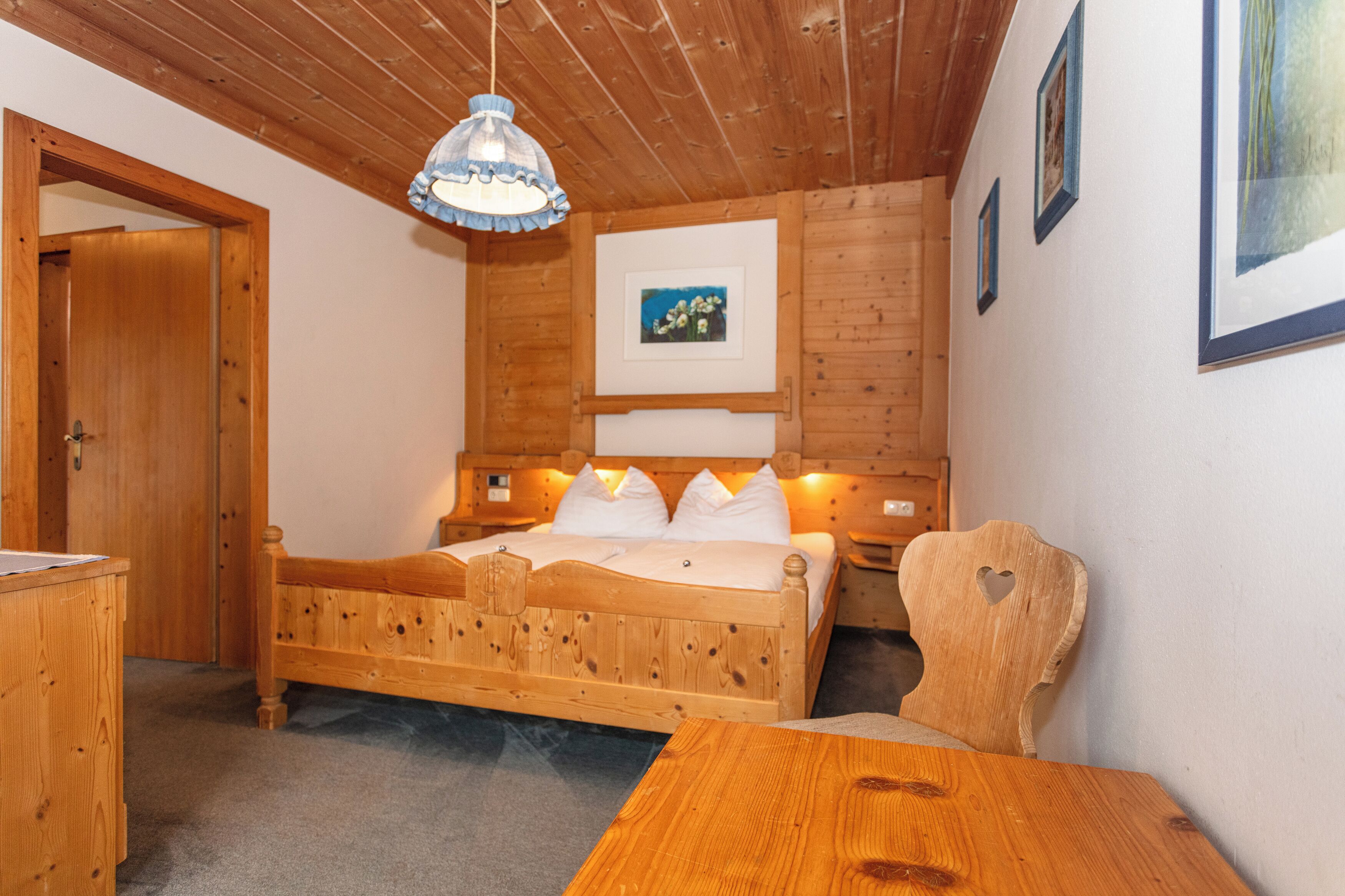 Ein hölzernes Bett mit weißer Bettwäsche, ein Nachttisch aus Holz, ein Holzstuhl und ein Schreibtisch aus Holz.