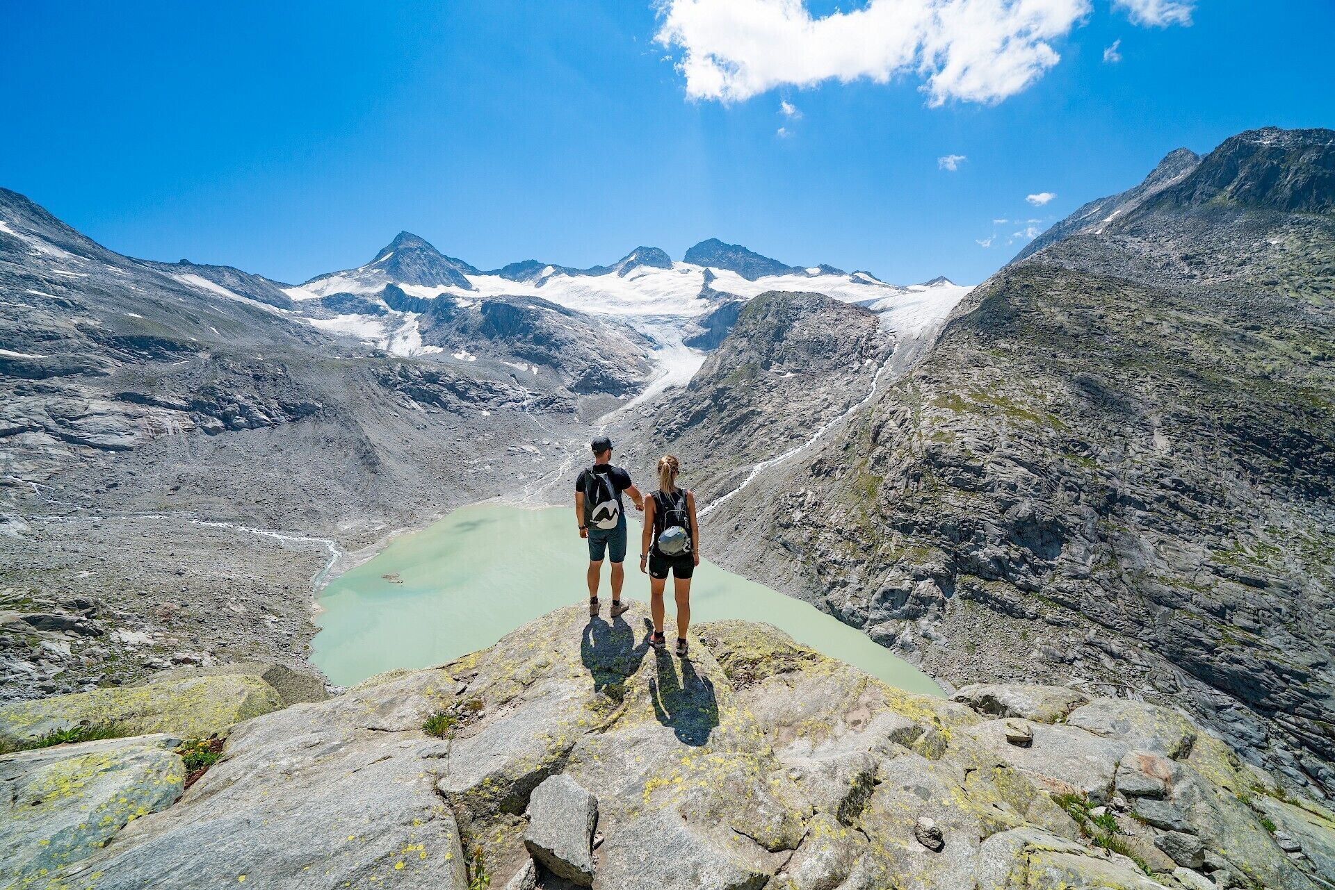 To personer står på en klippe med udsigt over en dal med en gletsjer og bjerge i baggrunden.