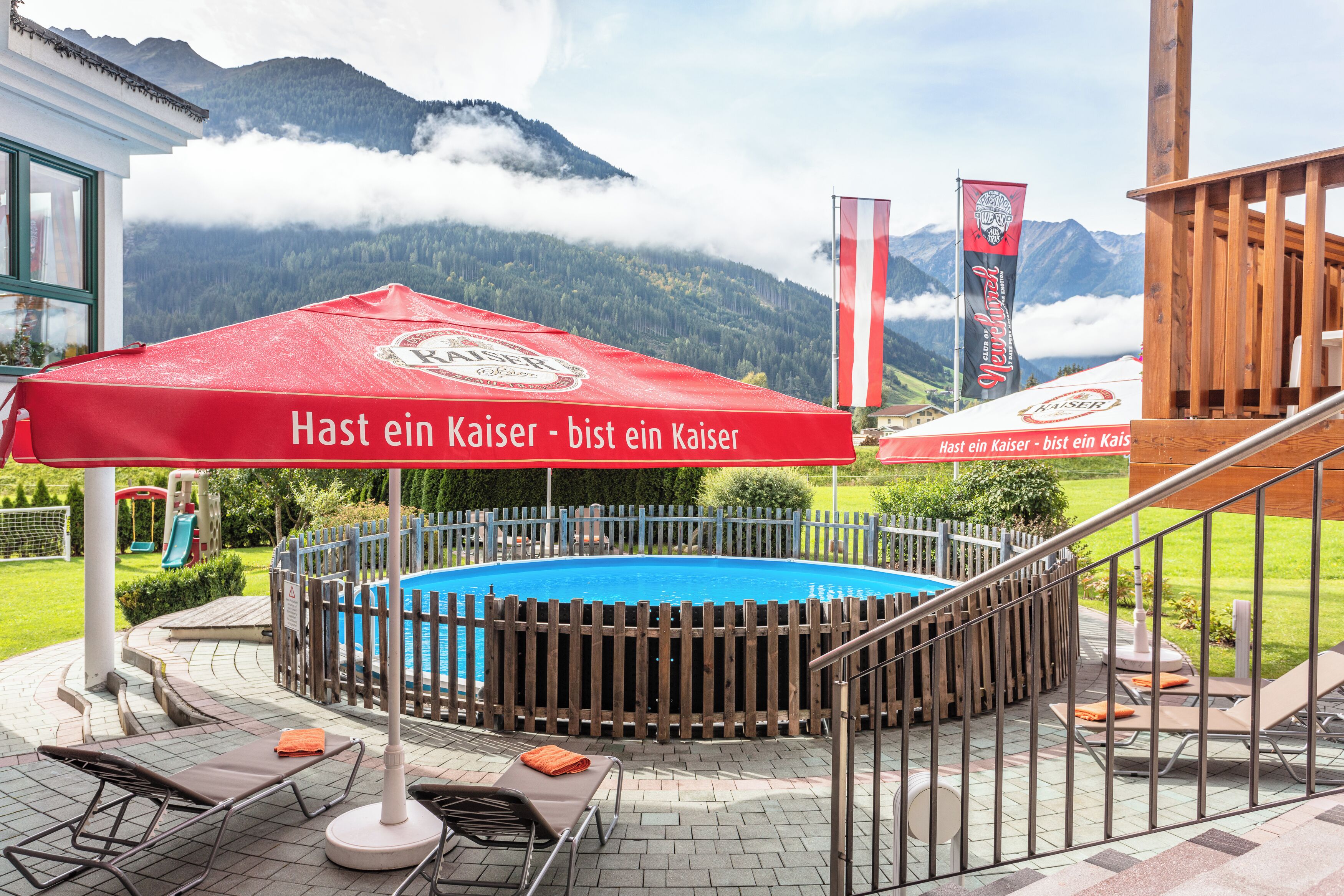 A swimming pool with a wooden fence, lounge chairs, and a red umbrella with text.