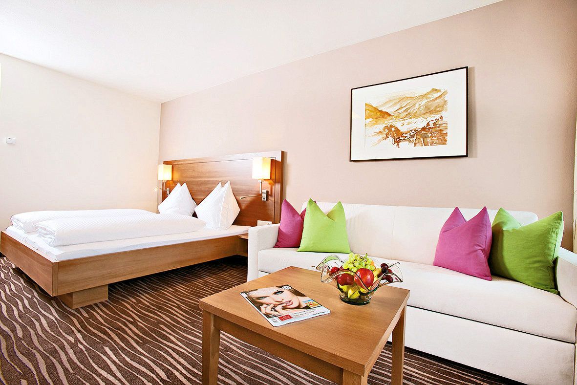 A hotel room with a bed, a sofa, a coffee table with a magazine, and a painting on the wall.