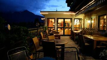 An outdoor patio area with tables and chairs, a view of a mountain, and a building with large windows.