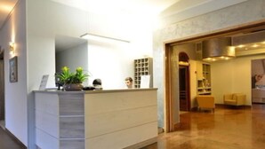 Un comptoir d’accueil agrémenté d’une plante, d’un miroir et d’une étagère où sont posés divers objets. Une porte donne accès à une pièce qui contient une chaise et une table.