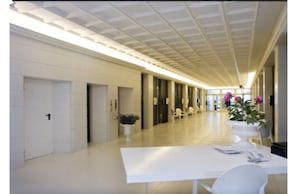 A hallway with white walls, a tiled floor, and a ceiling with recessed lighting. There are doors on the left, a table with chairs and a vase of flowers on the right, and a potted plant in the center. - Hotel Smeraldo (Abano Terme)