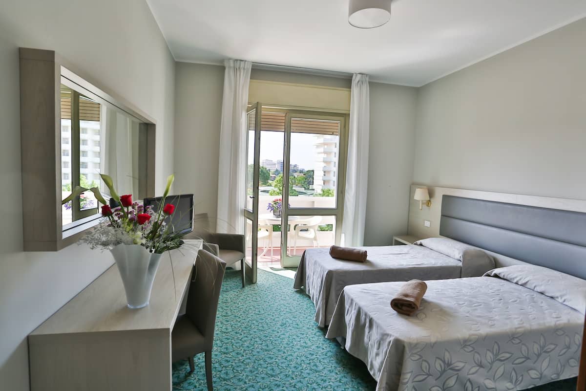 A hotel room with two beds, a desk, a chair, a vase with flowers, and a view of the outside.