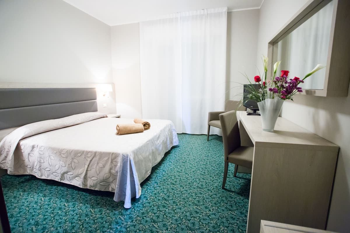 A hotel room with a bed, a desk, a chair, a vase with flowers, and a mirror.