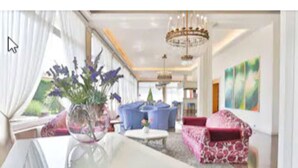 A spacious living room with a chandelier, a large window with curtains, a red patterned sofa, and a blue armchair. - Hotel Smeraldo (Abano Terme)