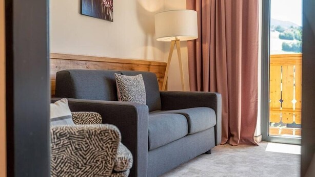 A modern living room with a grey sofa, patterned cushions, a floor lamp, and a view of a wooden deck through a window with curtains. - Zum Stadttor (Schladming)