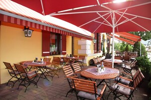 Un área de patio al aire libre con mesas redondas y sillas de madera, protegida por sombrillas rojas.