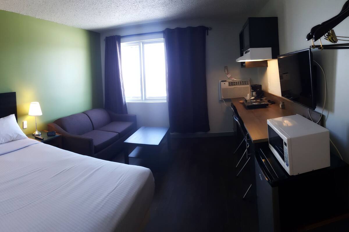 Habitación de hotel con cama, sofá, escritorio con computadora, lámpara y ventana con cortinas.