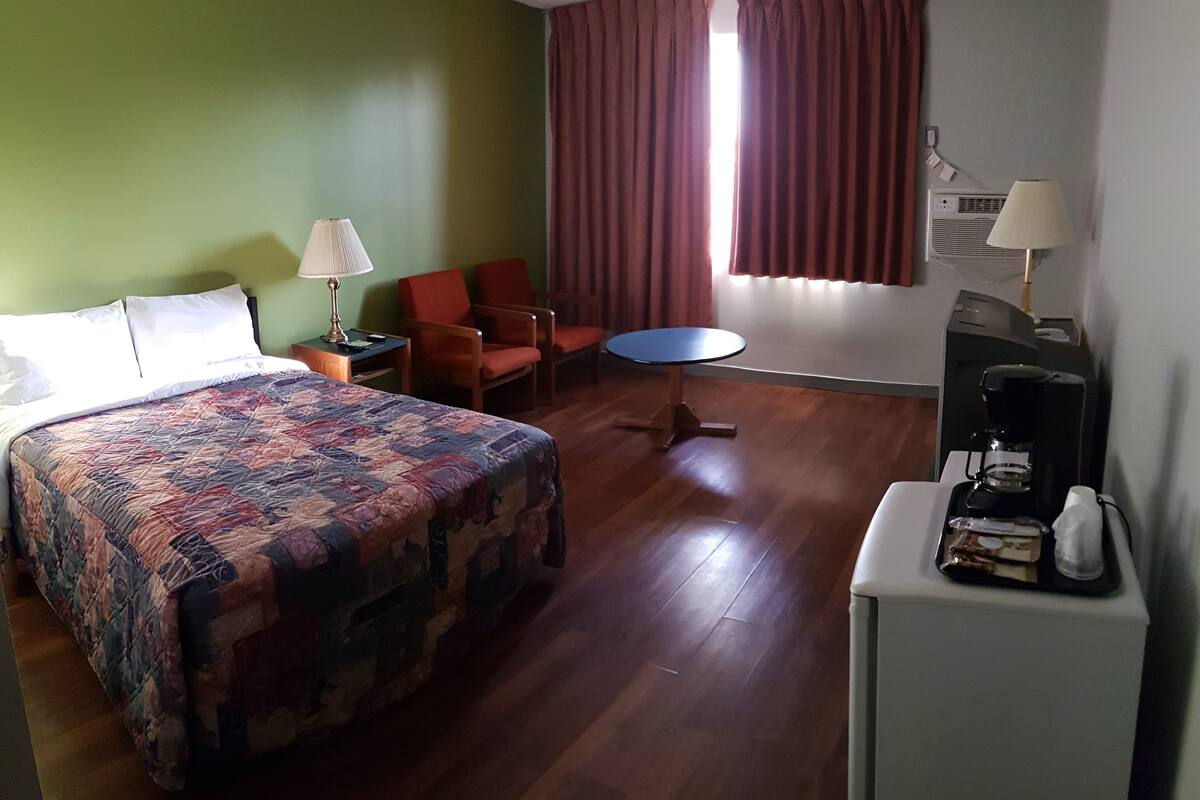 Una habitación de hotel con una cama grande, un escritorio con cafetera, dos sillas, una mesita redonda y una ventana con cortinas.