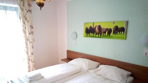 Una cama doble con sábanas blancas, cabecera de madera, un cuadro de ganado colgado en la pared y una ventana con cortinas florales.