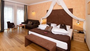 A hotel room with a large bed, a sofa, a small table, and a chair. There are two bedside lamps and a window with curtains. - Hotel Engel (Sluderno)