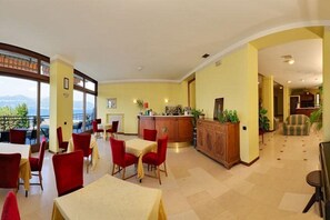 A hotel lobby with a dining area, a reception desk, and a view of the outdoors. - Europa (Ispra)