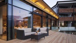 Un área de estar moderna al aire libre, con terraza de madera, paredes de vidrio y un comedor visible a través de las ventanas.