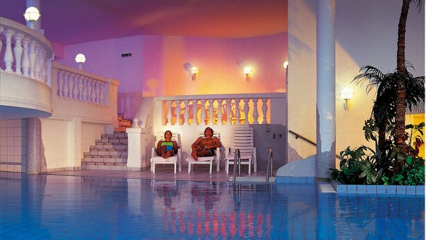 A pool area with a staircase leading to a balcony, lounge chairs, and a wall with decorative columns. - Hotel Alpina Resort (Wenns)