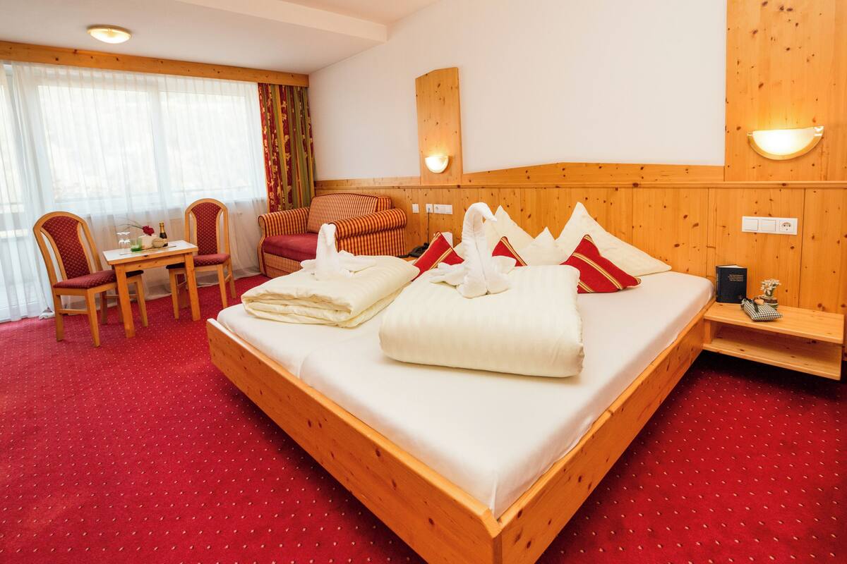 A hotel room with a large bed, a wooden headboard, a small table with chairs, and a window with curtains.